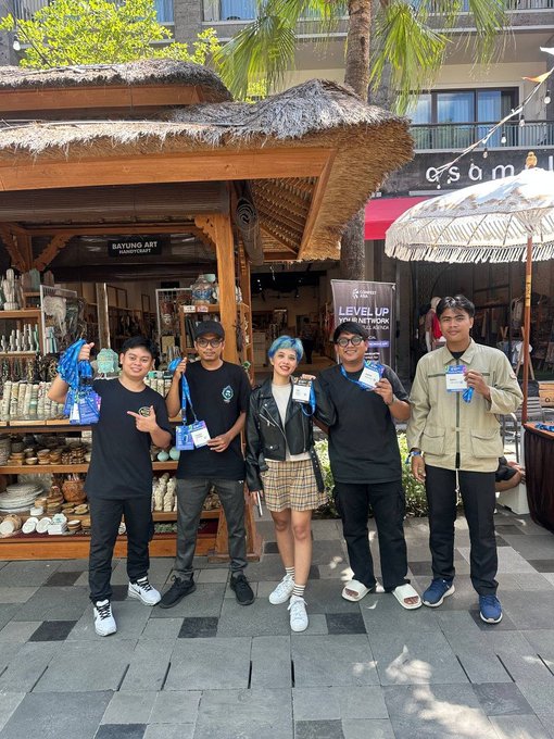 A group of five young people posing together at an outdoor venue, with some holding event badges, standing near a 'BAYUNG ART Handicraft' shop.