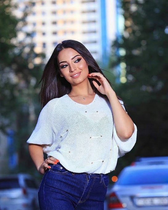 A young woman with long dark hair poses outdoors in a white sparkly top and blue jeans, with blurred city buildings and a car in the background.