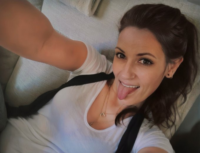 A smiling young woman with dark hair takes a playful selfie, sticking her tongue out while wearing a white t-shirt and delicate necklaces.