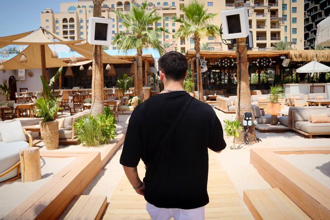 A man in a black t-shirt and white pants stands with his back to the camera on a wooden walkway at a luxurious beachside resort, with palm trees, outdoor lounge seating, and large speakers visible around him. The "GM" (Good Morning) caption fits the scene as a stylish greeting from an upscale tropical destination.