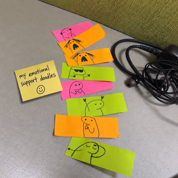 A handwritten yellow sticky note labeled "my emotional support doodles" sits beside a chain of colorful sticky notes (pink, orange, and green) featuring hand-drawn cartoon characters, with a pair of black earbuds resting nearby on a gray surface. The caption playfully connects to the Vietnamese post about hoping Binance features the user's "emotional support doodles," likening the chain of connected drawings to the cryptocurrencies mentioned being "taken too far" on their journey.