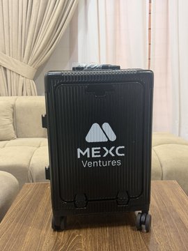 A black MEXC Ventures branded suitcase photographed on a wooden table in a living room setting.
