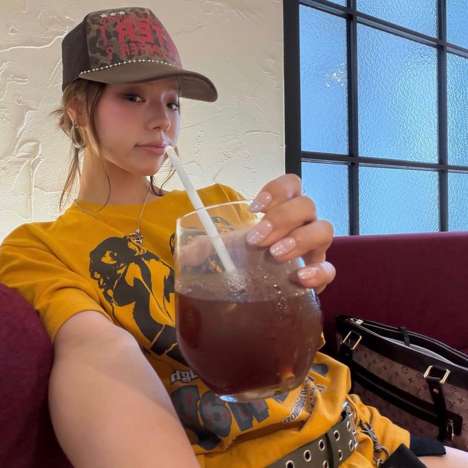 A young woman in a yellow graphic t-shirt and trucker hat drinking from a large glass with a straw in a cafe booth.