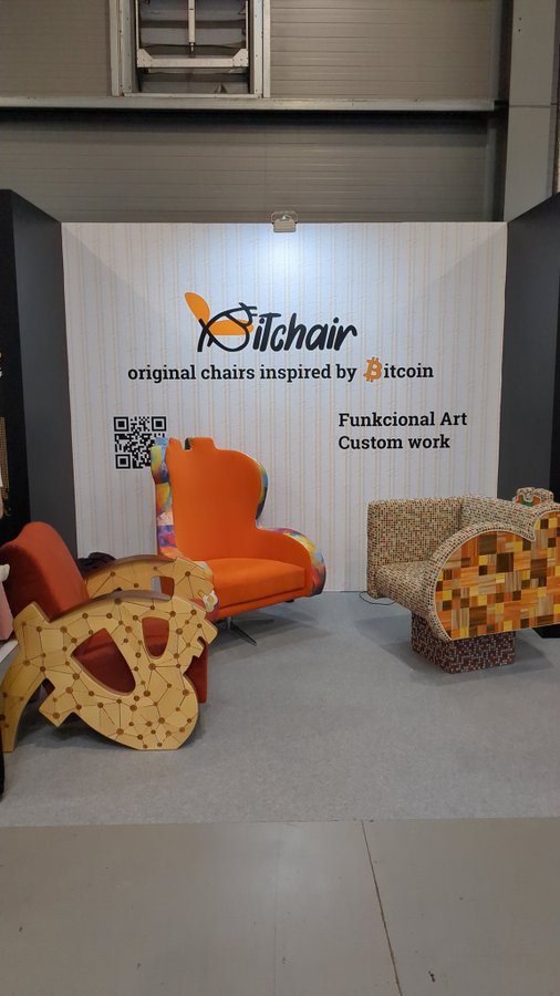 The image features two chairs displayed at an indoor exhibition or showroom. One of them has a unique design resembling a robot's head, while the other appears more traditional. The chairs are placed against a wall adorned with a sign that reads "If we had 2 pick 1 thing @BTCPrague worth looking at/ listening to.. sry Max Hillebrand / @saylor ... bulla was the steal of the century, but.. sure bauhaus, chippendale, eames, egg etc designs were/ r cool". The chairs are positioned in front of a display case or booth that showcases other furniture items.