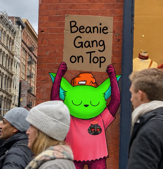 A green cartoon character wearing a pink shirt and orange beanie holds up a cardboard sign reading "Beanie Gang on Top" on a city brick wall, while pedestrians walk past. The mural aligns with the post's shoutout to @BeanieGangg, visually representing the community's signature beanie-wearing mascot and rallying slogan.