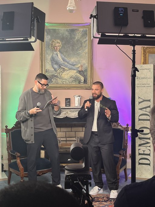 Two men stand on a stage holding microphones during a 'DEMO DAY' event, with a Castitec banner, professional lighting, and a portrait painting visible in the background.