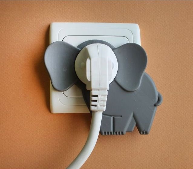 A gray elephant-shaped child safety cover is plugged into a white electrical outlet mounted on a peach-colored wall, with a white cord extending downward.