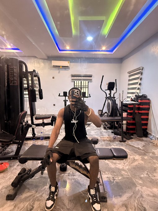 A selfie taken in a home gym with colorful LED ceiling lights, showing a muscular person wearing a black sleeveless shirt, shorts, gloves, and an Apple Watch, sitting on a weight bench while holding a phone to take the photo.