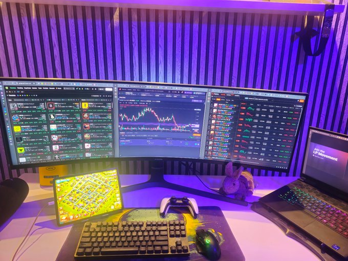 A multi-monitor trading setup featuring three large displays running FabriqTrade, GMGN, and MeteoraAG for tracking crypto pools and LP positions, alongside a laptop showing a Clash of Clans game screen, a game controller, a mechanical keyboard, and a small plush bunny on the desk under purple ambient lighting.