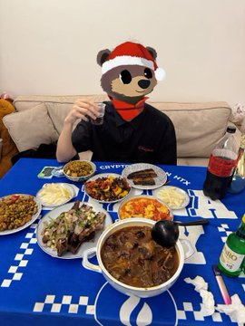 A person wearing a teddy bear head mask and a Santa hat sits at a table covered with a blue soccer-themed cloth and various dishes of food.