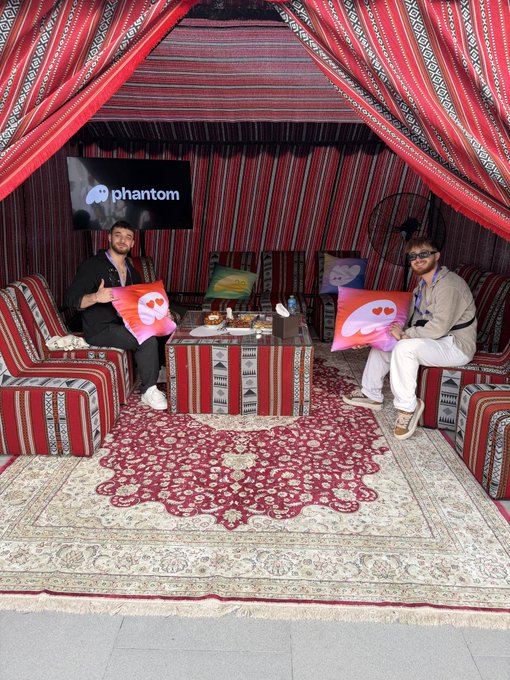 Two men smile while seated in a tent-like lounge with red and black striped fabrics, each holding a pink "Phantom" branded pillow, with a TV screen displaying the "Phantom" logo behind them, fitting the cheerful "GM" (Good Morning) greeting.