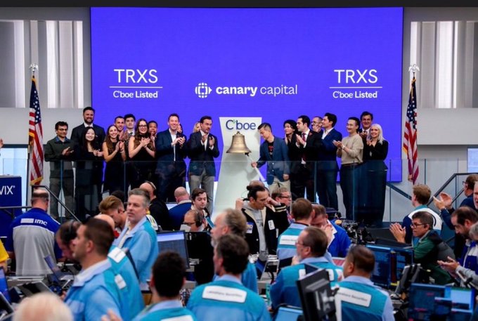 A group of people celebrate on a podium at the Cboe stock exchange, clapping near a bell, with the background screens displaying 'TRXS Cboe Listed' and 'canary capital'.