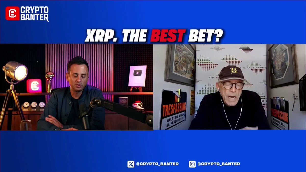 The image features a man sitting at a desk with a microphone, likely recording an interview or podcast discussion about Bitcoin. The background of the image shows a blue banner that reads "XRP. THE BEST BEAT?" along with some text. There are two men in the image - one is seated behind the other who appears to be talking on the phone while the first man sits at his desk, possibly preparing for or just finished an interview or podcast recording about Bitcoin and XRP.