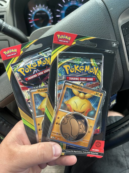 A hand holds two Pokémon Trading Card Game blister packs featuring Makuhita, taken inside a car with a steering wheel and dashboard gauges visible in the background. The caption "Bought for the cosmos And maybe a meowth?" humorously reflects on the uncertainty of what cards might be found inside, as Meowth is a fan-favorite Pokémon often hoped for in booster packs.