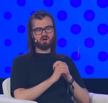 The image shows a man with long brown hair and a beard, wearing glasses and a black t-shirt, seated on a white couch against a blue background with purple polka dots, appearing to speak at a panel or interview. There is no visible text, numbers, or tickers in the image. However, the context provided suggests this is Murad, a figure known in the crypto community for his memecoin recommendations, with the caption likely prompting a retrospective on the performance of those previously endorsed tokens two years later.