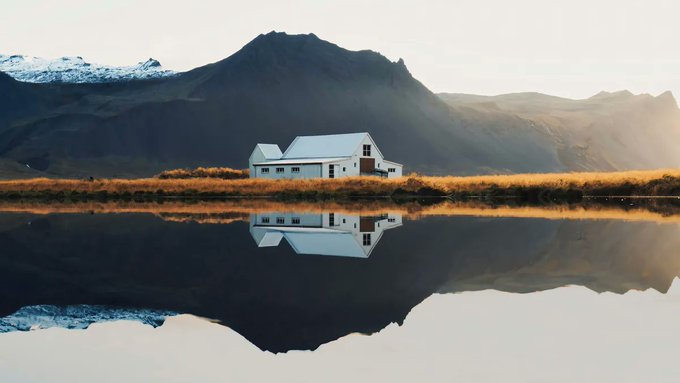 A serene landscape features a white house and snow-capped mountains perfectly reflected in the calm waters of a lake under a bright sky.