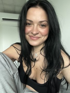The image features a woman with long black hair wearing a black bra top or bikini bottom. She has her arms around herself as she poses for the camera. The background of the photo shows a white wall, which contrasts nicely with the woman's dark hair. There is also an air conditioning unit visible in the background.
