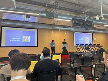 A presentation is taking place in a modern conference room with two presenters at the front, and a screen displaying the text 'slido.com' and '#5883 SUB' alongside a QR code.