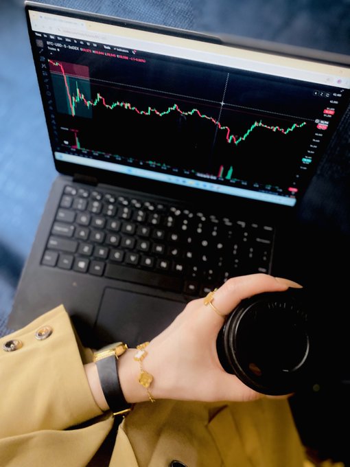 A person's hand adorned with gold bracelets rests on a coffee cup beside a laptop displaying a stock or crypto trading chart.