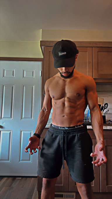 A shirtless, muscular man wearing a black 'ADID' cap and black shorts poses in a kitchen, showing off his toned physique with arms extended slightly outward.