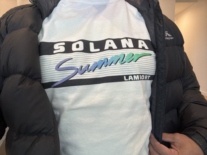 The image features a person wearing a white t-shirt with the word "solana" printed across the front, along with the phrase "Summer Lamiotart". The shirt has a hoodie or puffy jacket draped over it, suggesting that the weather might be cool or rainy outside. The person is standing against a wall and appears to be indoors.