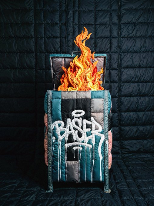 An artistic photograph of a teal and black puffer jacket with graffiti-style text reading "BASER" and an orange/red flame sculpture emerging from inside it, set against a dark quilted background.