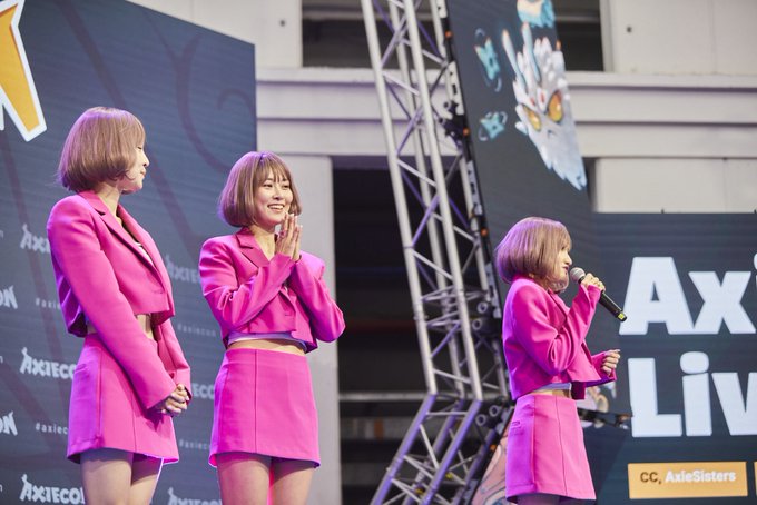 Three women in matching pink outfits perform on stage at an Axie Live event, with one holding a microphone.