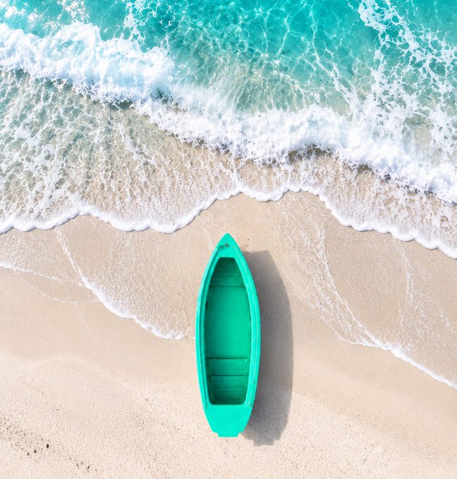 An aerial view of a small green boat resting on a sandy beach as turquoise waves wash ashore.