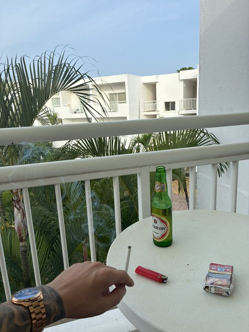 A first-person view from a balcony holding a lit cigarette, with a Budweiser beer bottle, lighter, and cigarette pack on a white table, overlooking a tropical resort with palm trees.