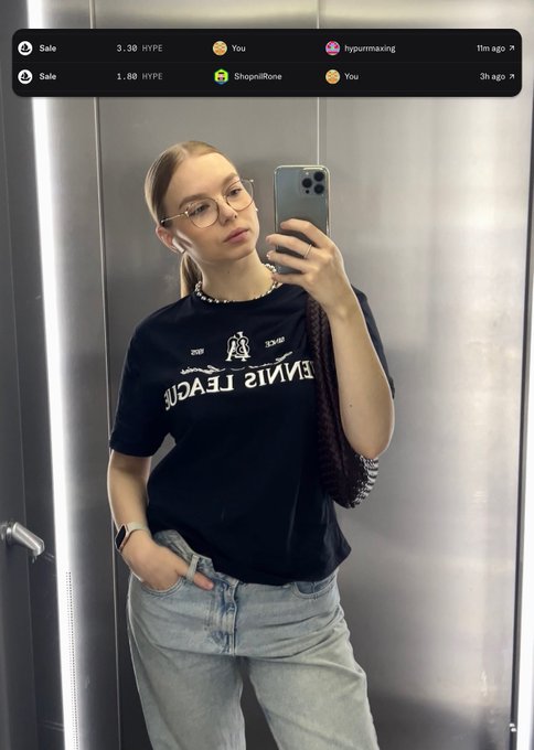 A young woman takes a mirror selfie wearing a black graphic t-shirt (with text appearing as 'FINA JEACN'), light blue jeans, glasses, and a pearl necklace. Overlay text shows NFT transactions: 'Sale 3.30 HYPE hyurmwaxing' and 'Sale 1.80 HYPE ShopnilRone'.