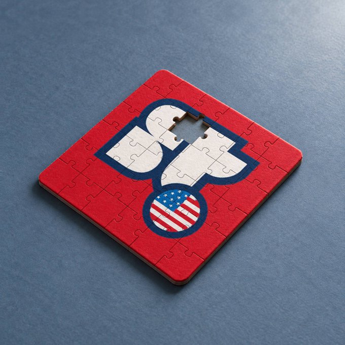 A square jigsaw puzzle rests on a gray surface, featuring a red background with a white Spartan helmet silhouette containing the American flag on its crest, with one puzzle piece missing.