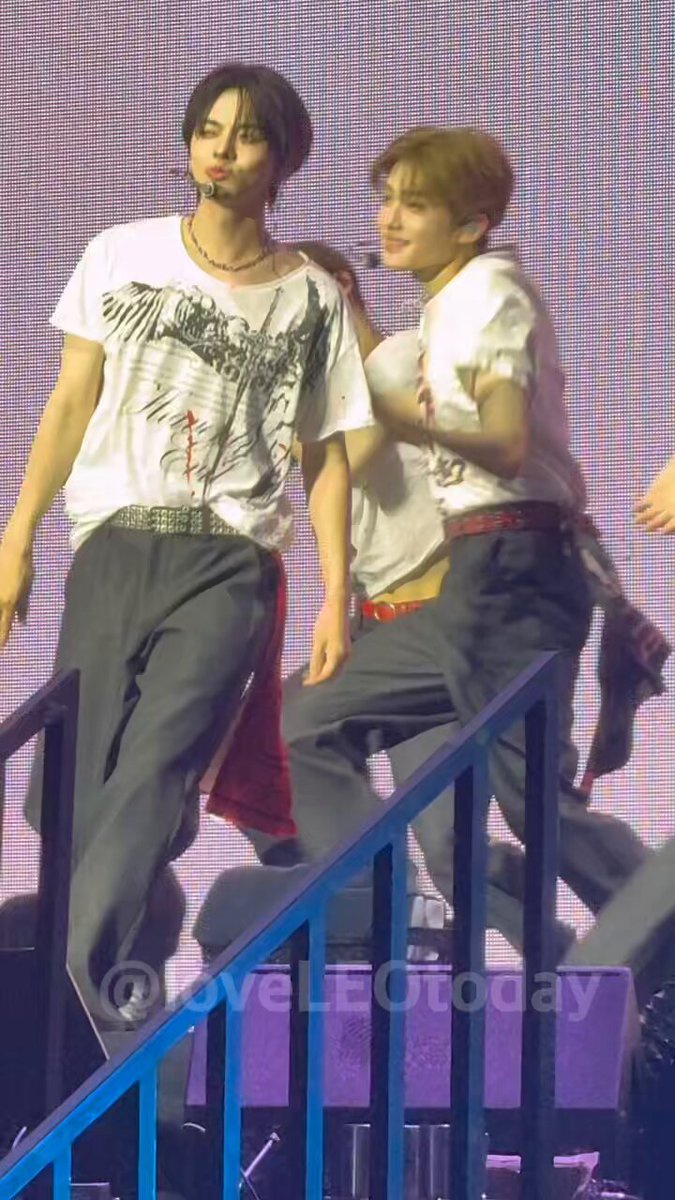 The image features two people standing next to each other, possibly at a concert or event. They are both wearing t-shirts with various designs. The person on the left has their arm around the other person's shoulder, creating an intimate atmosphere between them. In the background, there is a stage set up for the event, and a caption can be seen below the image that reads "260614 OMG 파드라이브원 #ALD1 #알파드라이브원 #LEO #리오".