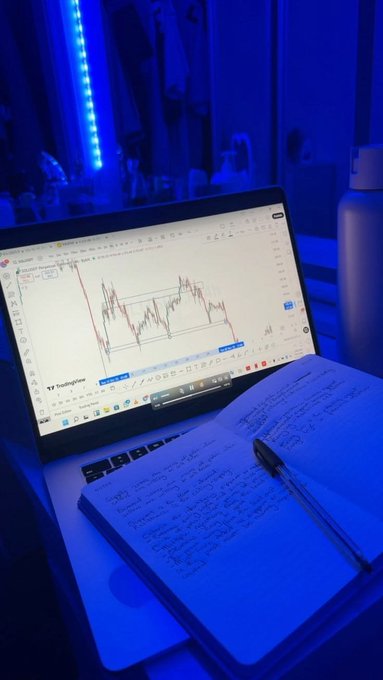 A trading setup featuring a laptop displaying a GBP/USD chart on TradingView alongside a handwritten notebook and pen, bathed in moody blue lighting—capturing the discipline of journaling and technical analysis during a bull market, reminding traders to keep learning and stay patient even when the charts turn red.