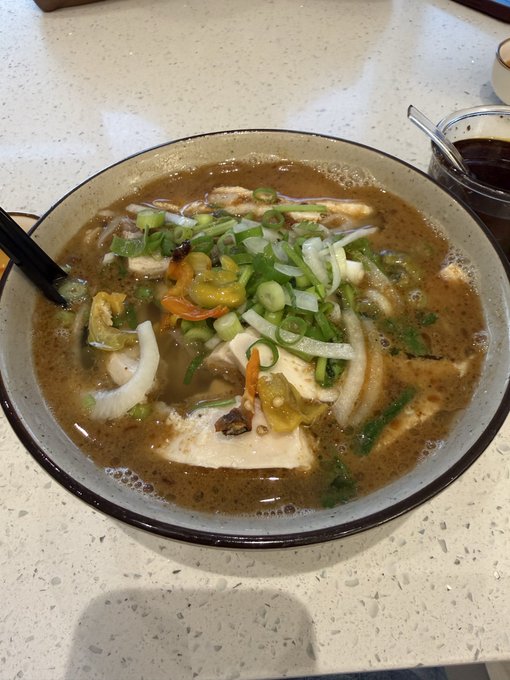 Close-up photo of a bowl of Asian soup or noodle dish garnished with green onions, containing tofu, mushrooms, and vegetables in a brown broth, with a black spoon resting in the bowl on a speckled countertop.