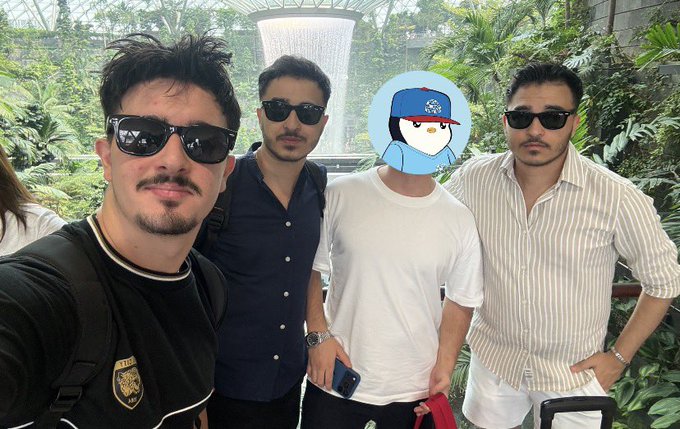 Four men pose for a selfie at what appears to be Jewel Changi, standing in front of the HSBC Rain Vortex indoor waterfall and tropical garden, with one person's face covered by a cartoon owl sticker.