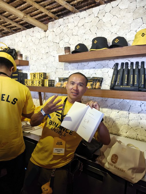A man wearing a yellow 'BINANCE ANGELS' shirt and lanyard holds an open notebook while standing in a merchandise shop with Binance products on display.