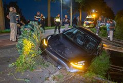 A car rests in a roadside ditch at night with debris scattered around, while bystanders and an emergency vehicle are visible in the background.