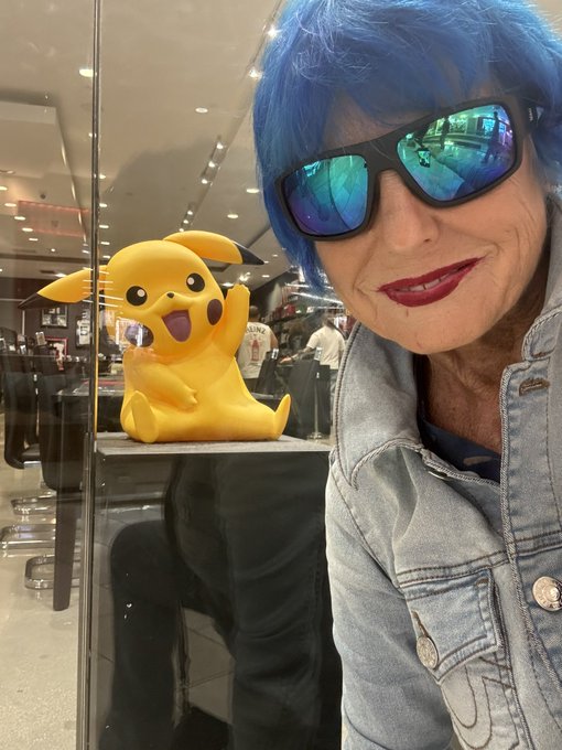 A selfie of a person wearing a blue wig, reflective sunglasses, and a denim jacket, posing next to a Pikachu statue visible behind glass.