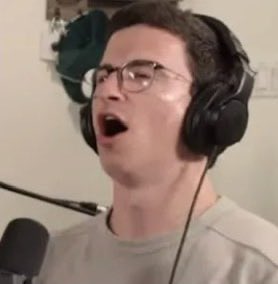 A young man wearing glasses and headphones is captured mid-yell with his mouth wide open, positioned in front of a microphone as if broadcasting or reacting to something. His exaggerated, almost ecstatic expression visually amplifies the post's caption "Shorts got liquidated," conveying the chaotic excitement of a financial squeeze.