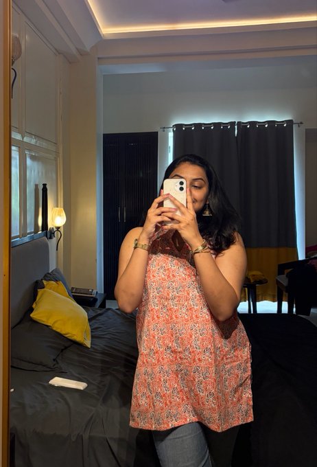 A mirror selfie of a woman wearing a sleeveless orange patterned top in a bedroom, taking the photo with her phone partially covering her face.