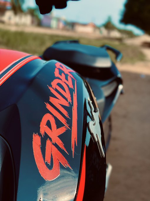 A close-up shot of a motorcycle's dark blue tank displays the word "GRINDER" in red graffiti-style lettering with red trim accents.