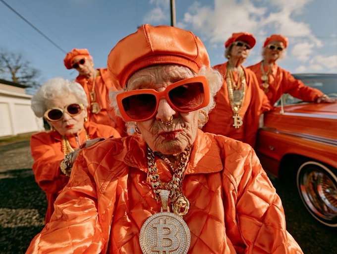 A group of elderly women dressed in matching orange outfits, hats, and sunglasses pose outdoors, with the central figure wearing a large necklace featuring a Bitcoin 'B' logo, next to an orange car.