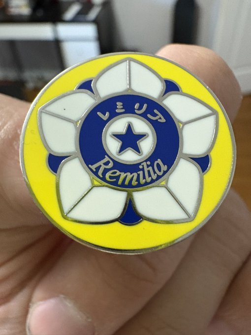 A close-up of a circular enamel pin held in a hand, featuring a white flower with a blue center containing a star and the text 'レミリア' and 'Remilia'.