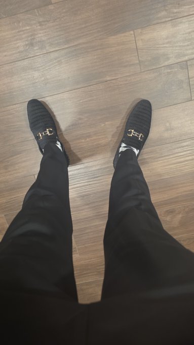 A person wearing black pants and black loafers with a gold horsebit detail stands on a wooden plank floor.