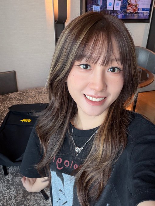 A smiling young woman with long brown hair and highlights takes a selfie wearing a black t-shirt with "pearlo" text visible and a silver necklace, with a TV displaying what appears to be a crypto/Binance-themed show in the background. The image appears to be a travel selfie capturing her experience in Vietnam, aligning with the post about the country's high crypto acceptance and paying for a SIM card with Binance via @pearbinance.
