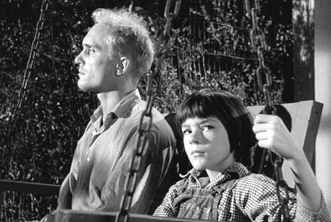 A black and white film still of an older man and a young boy sitting together on a swing set, looking off to the side.