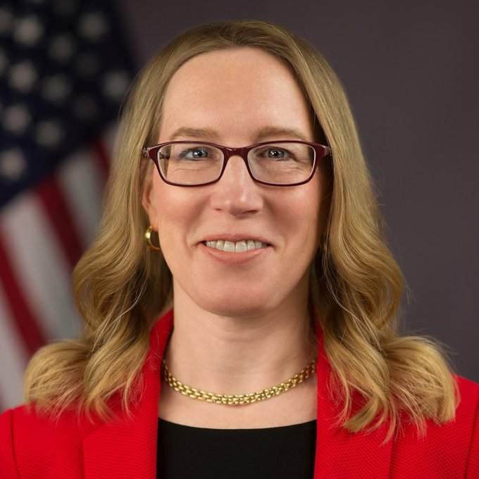 A professional headshot features a smiling woman with blonde hair and dark-rimmed glasses, wearing a red blazer and gold chain necklace, posed in front of an American flag. The formal portrait appears to be an official government or corporate photograph, though its specific connection to the post about ZIG Markets and onchain finance is unclear since no identifying text or names are visible in the image.