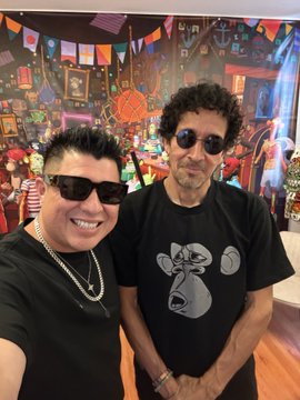 Two men posing together, one wearing a black shirt with a cartoon face design and the other in a black t-shirt, both wearing sunglasses in front of colorful papel picado decorations.