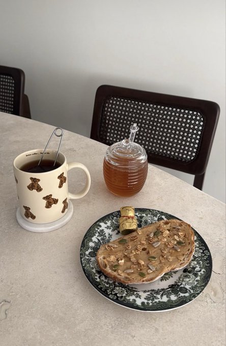 A breakfast scene on a marble table features a teddy bear-patterned mug of tea, a glass jar of honey, and a decorative plate with toast topped with seeds and pumpkin pieces.