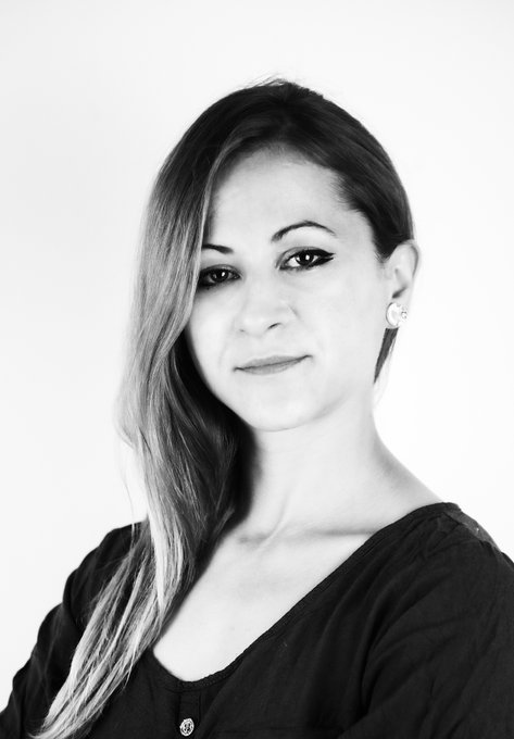 A black and white portrait photo of a woman with long dark hair and eyeliner, wearing a dark top, smiling softly at the camera.